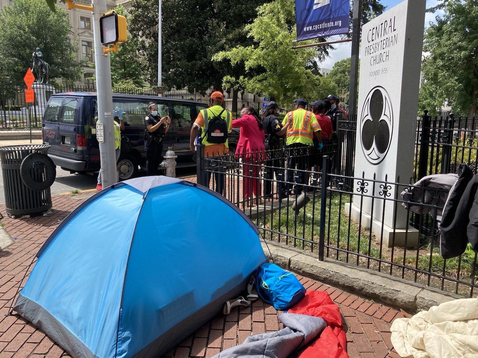 A blue tent sits on a sidewalk, and people in neon vests stand beside a van in the background.