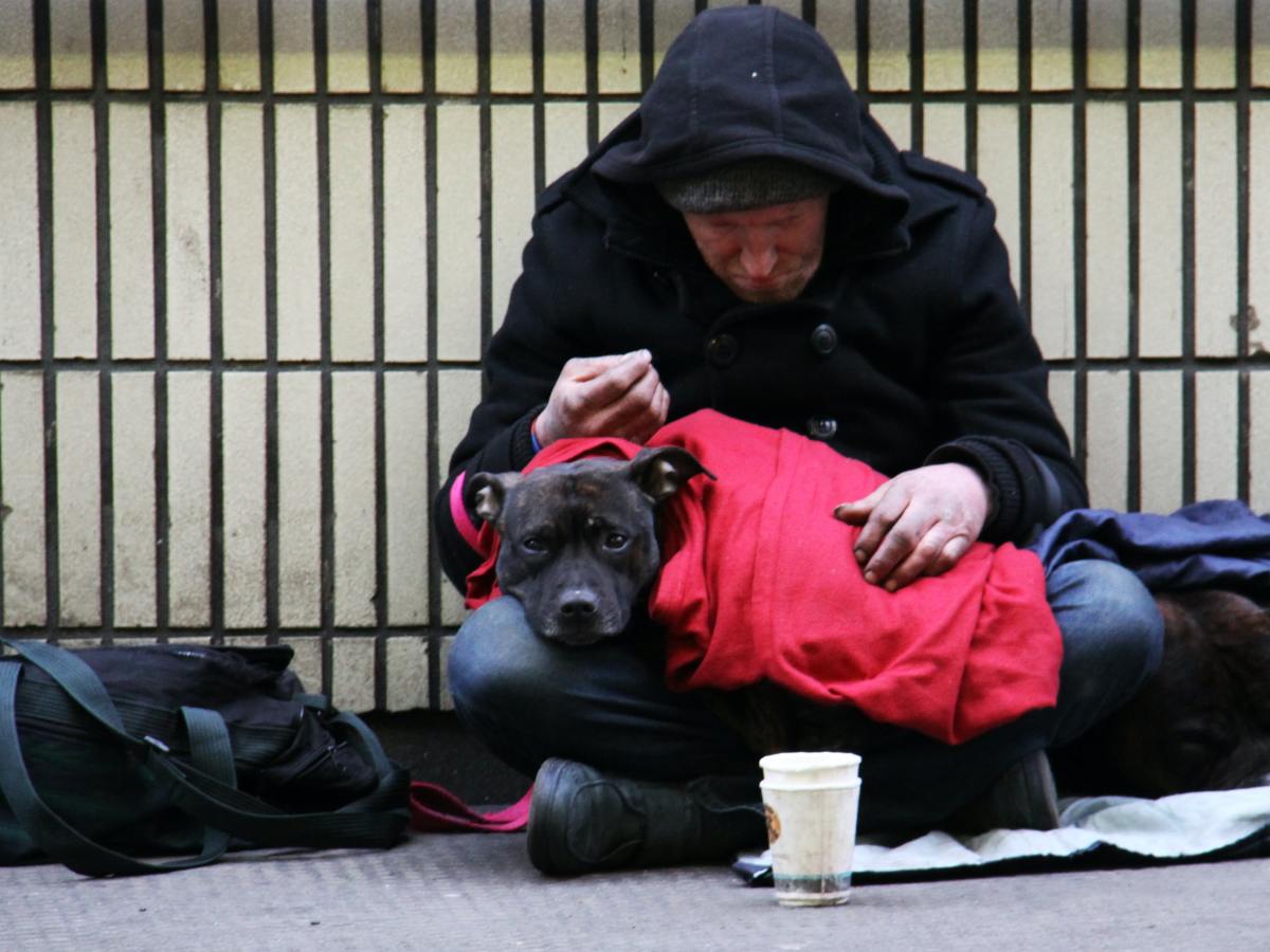 A homeless man sits on the sidewalk with dog in lap.