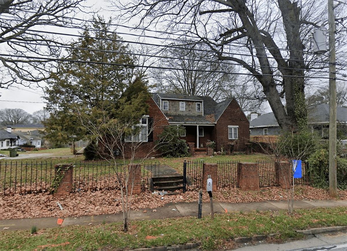 A small brown house with a gabled roof sits in the shade of old trees, surrounded by a green lawn.