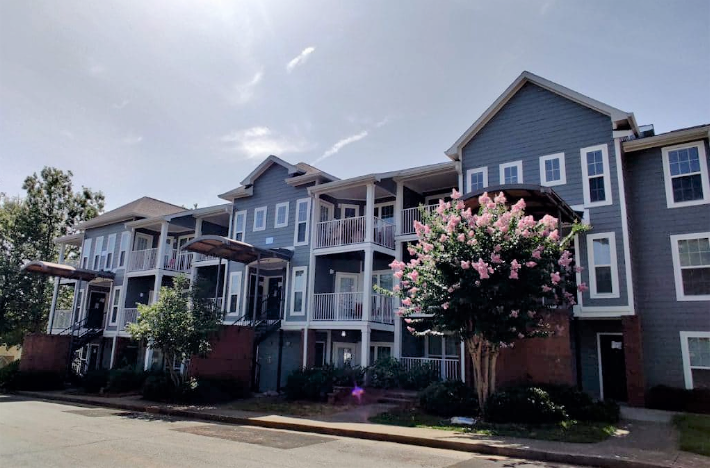 A pale blue, low-rise apartment building, with flowering trees shading a sidewalk.