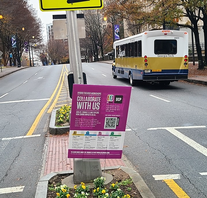 A pink sign soliciting community input for the zoning code rewrite is stuck into a median on a city street.