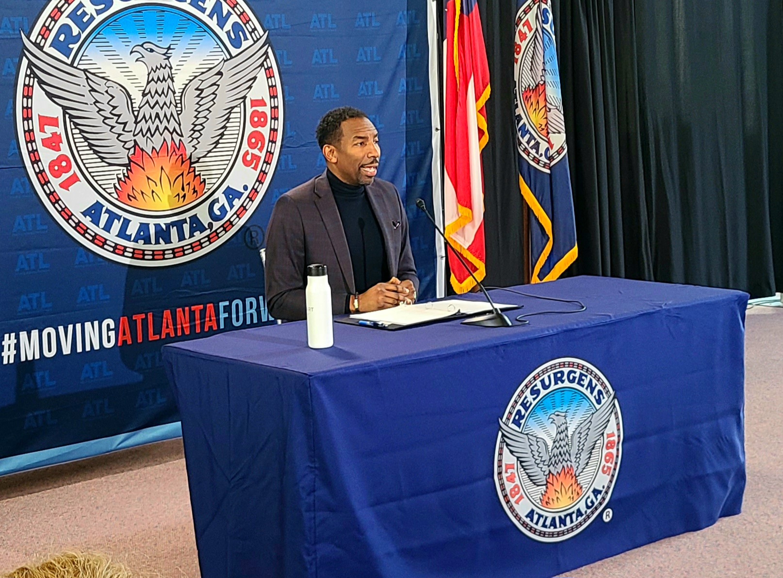 Andre Dickens, in a gray jacket and black shirt, sits behind a blue table emblazoned with the city's seal.