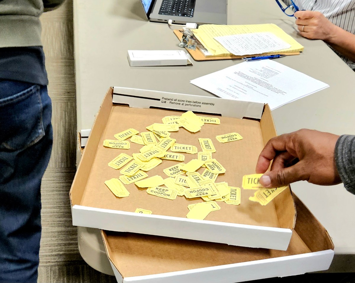 A cardboard box shaped like a pizza box holds dozens of yellow ticket stubs. A hand is dropping two more in.