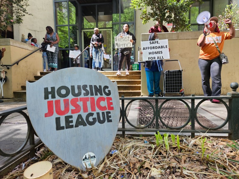 In the foreground, a wooden shield is emblazoned with "Housing Justice League." In the background, activists protest outside an office building, holding megaphones and signs.