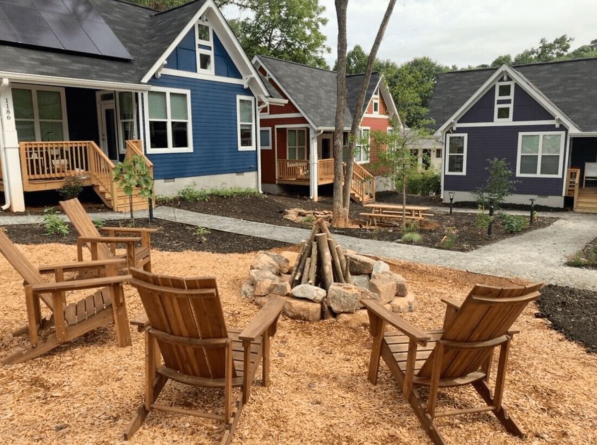 A photo of a campfire surrounded by wooden chairs. In the background are three colorful tiny homes.