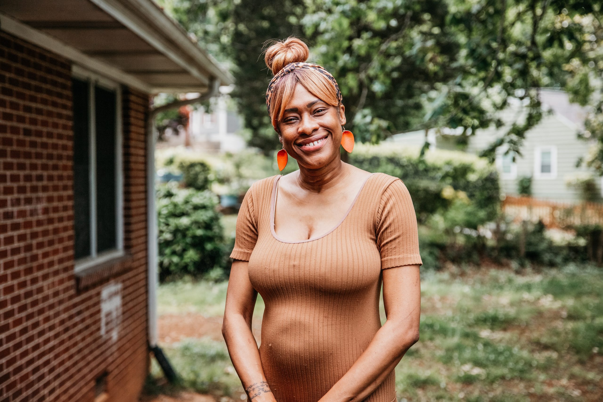 Shenika Phillips smiles bright while standing outside. She's wearing a short-sleeved brown jumpsuit and orange leaf-shaped earrings. Her hair is tied up in a bun. 