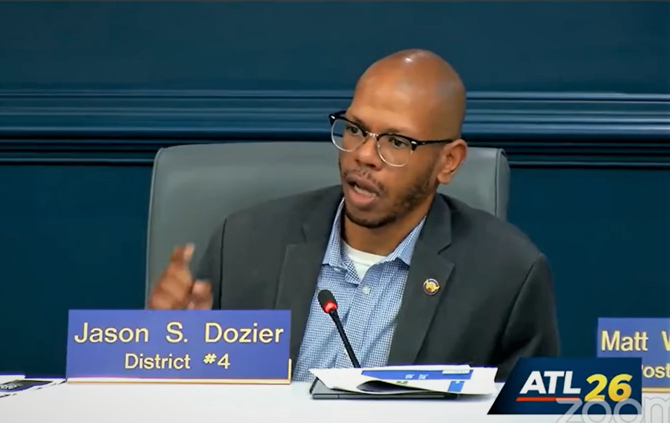 A screenshot of a stream of the Atlanta City Council's CD/HS Committee meeting, framing Councilmember Jason Dozier. A nametag is in front of him, and he's pointing his finger while speaking into a microphone.