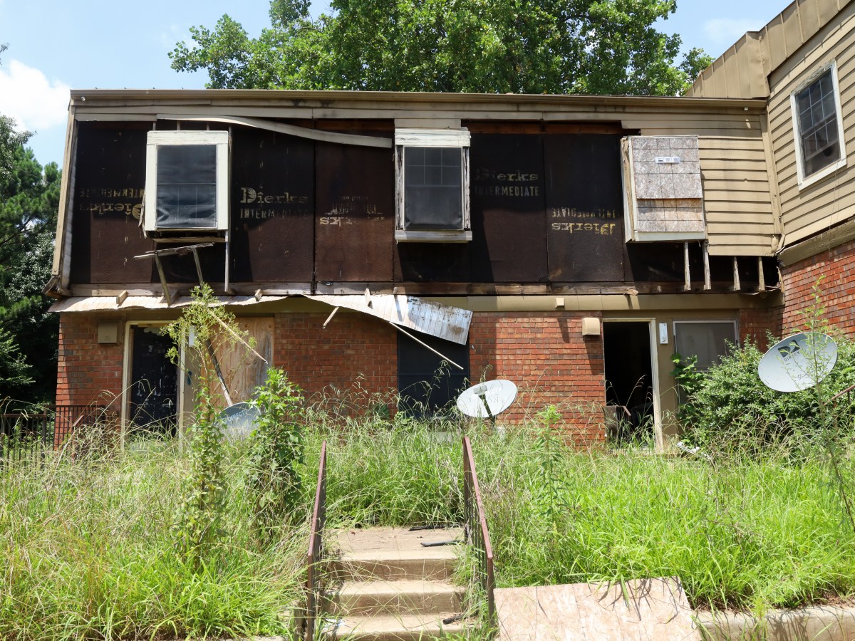 A photograph of a low-rise apartment building whose siding has all but fallen off. Weeds grow tall in the foreground.