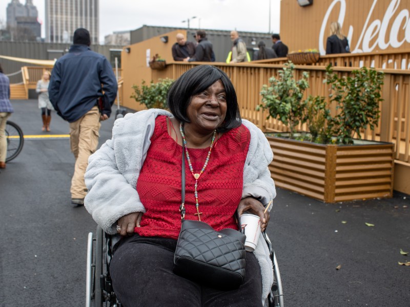A photo of an older Black woman in a wheelchair wearing a red shirt and white jacket. She's smiling, looking off camera.