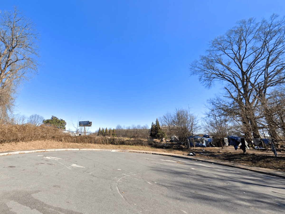 A picture of a vacant, overgrown lot, with a small tent encampment beneath trees to the right.