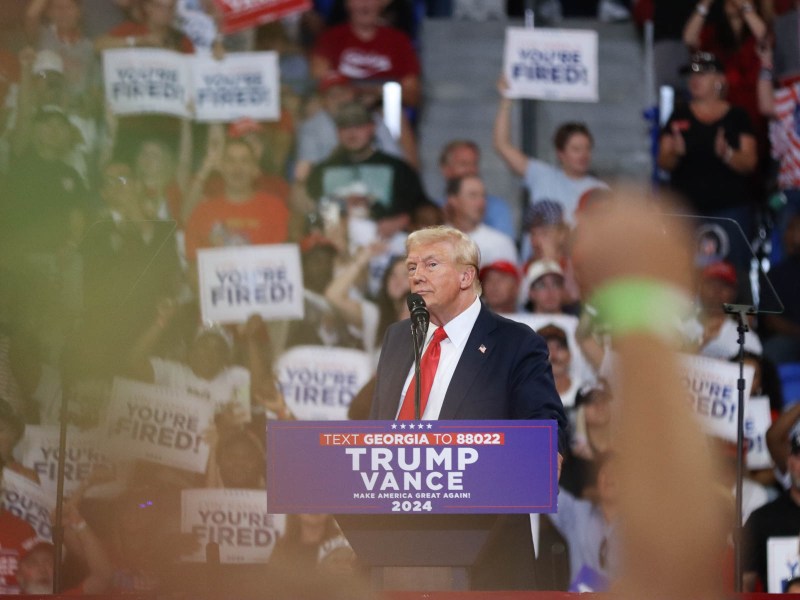 A photo of Donald Trump, in his usual red tie and dark suit, speaking before a crowd.