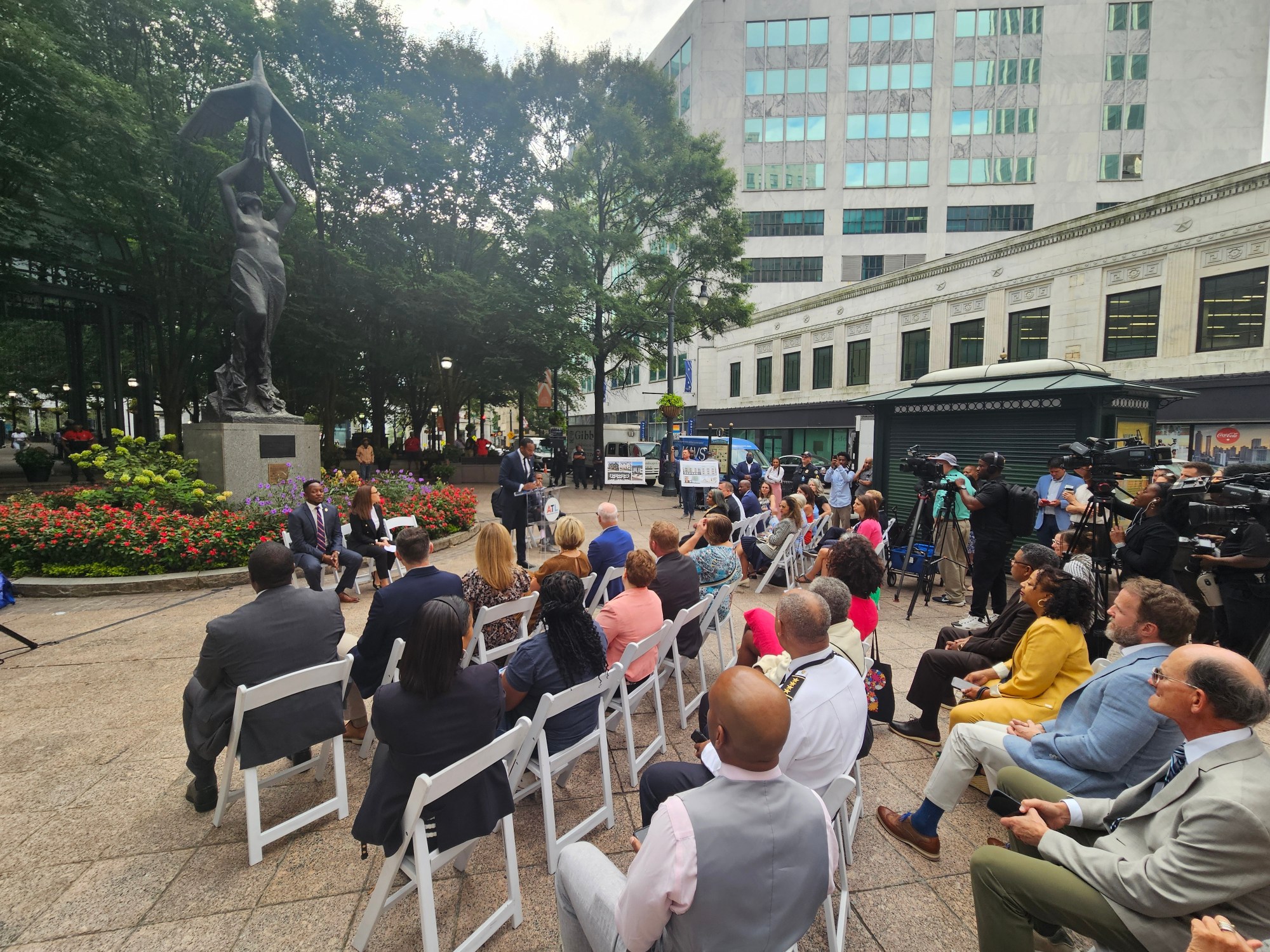 A photograph of a crowd assembled to watch Mayor Andre Dickens, seated under a bronze statue, make an announcement.