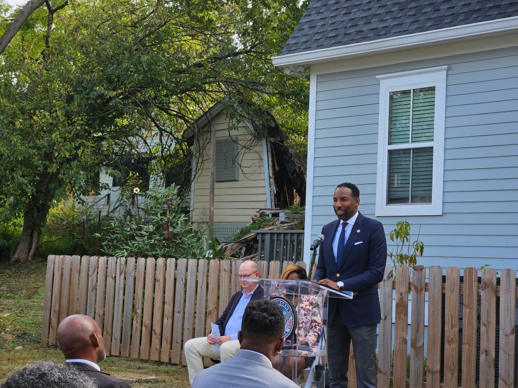 A photo of Andre Dickens speaking from a podium in front of a newly renovated house and an old dilapidated house.
