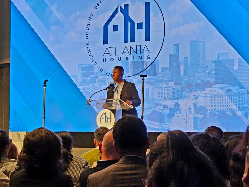 Terri Lee, wearing a dark suit and light blue blouse, speaks at a podium on a stage.