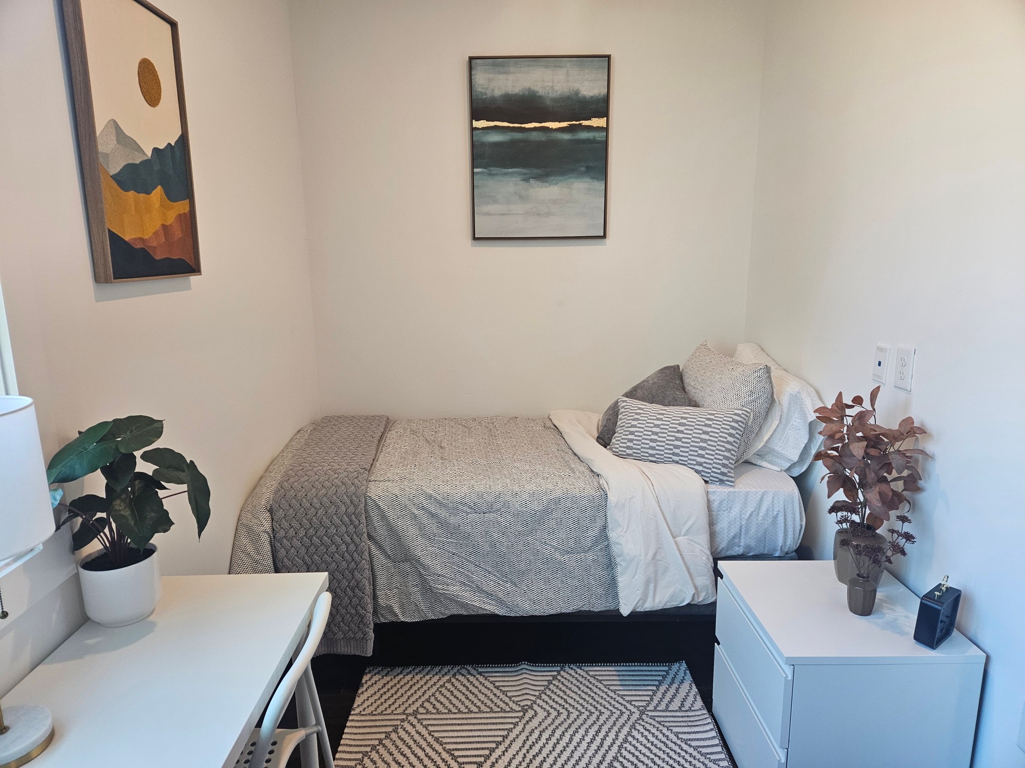 A photo of a small bedroom, with white walls and grey furniture and bedding.