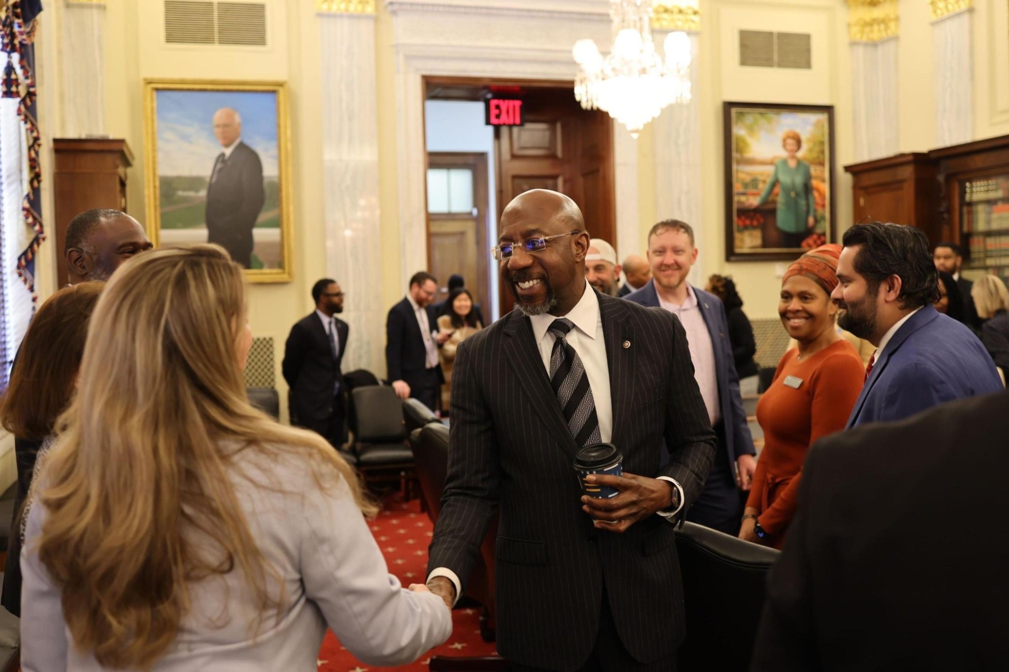 A photo of Raphael Warnock, wearing a black suit, shaking hands with a woman.