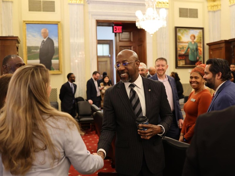 A photo of Raphael Warnock, wearing a black suit, shaking hands with a woman.