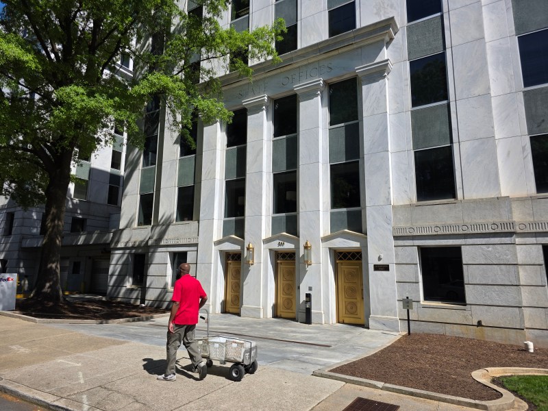 A photo of a state government office building downtown, its face marbled white. A man pushes a cart past it.