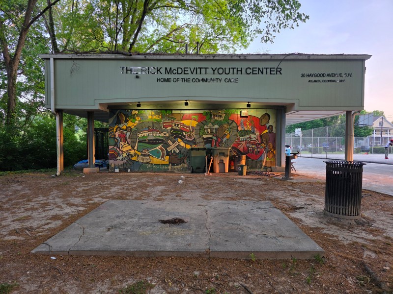 A photo of Atlanta’s aging Rick McDevitt Youth Center, a pale-green low-rise building featuring a colorful mural. The grounds are covered in pinestraw. Children play basketball in the background.