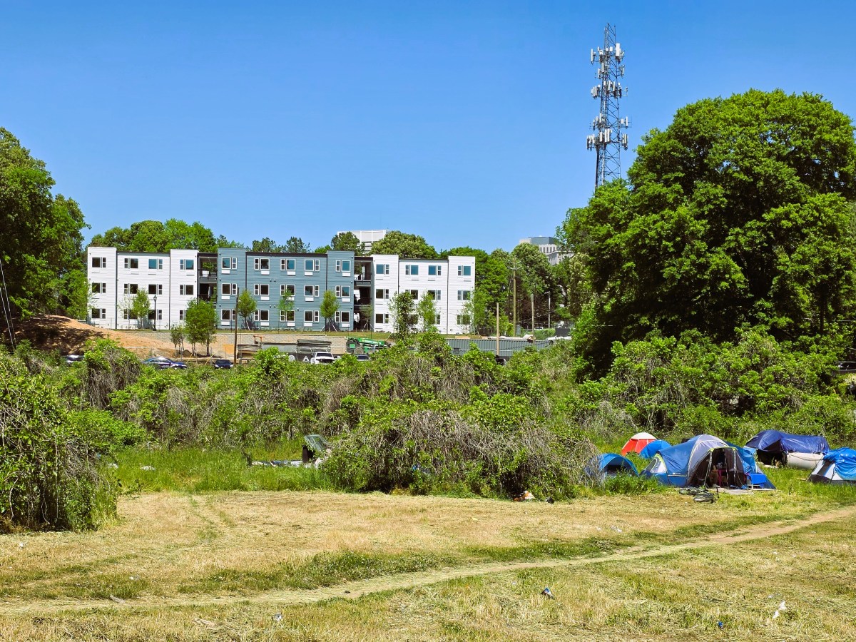 A grey and white apartment complex sits atop a hill. In the foreground, an overgrown lot is dotted with tents, where unhoused people live.