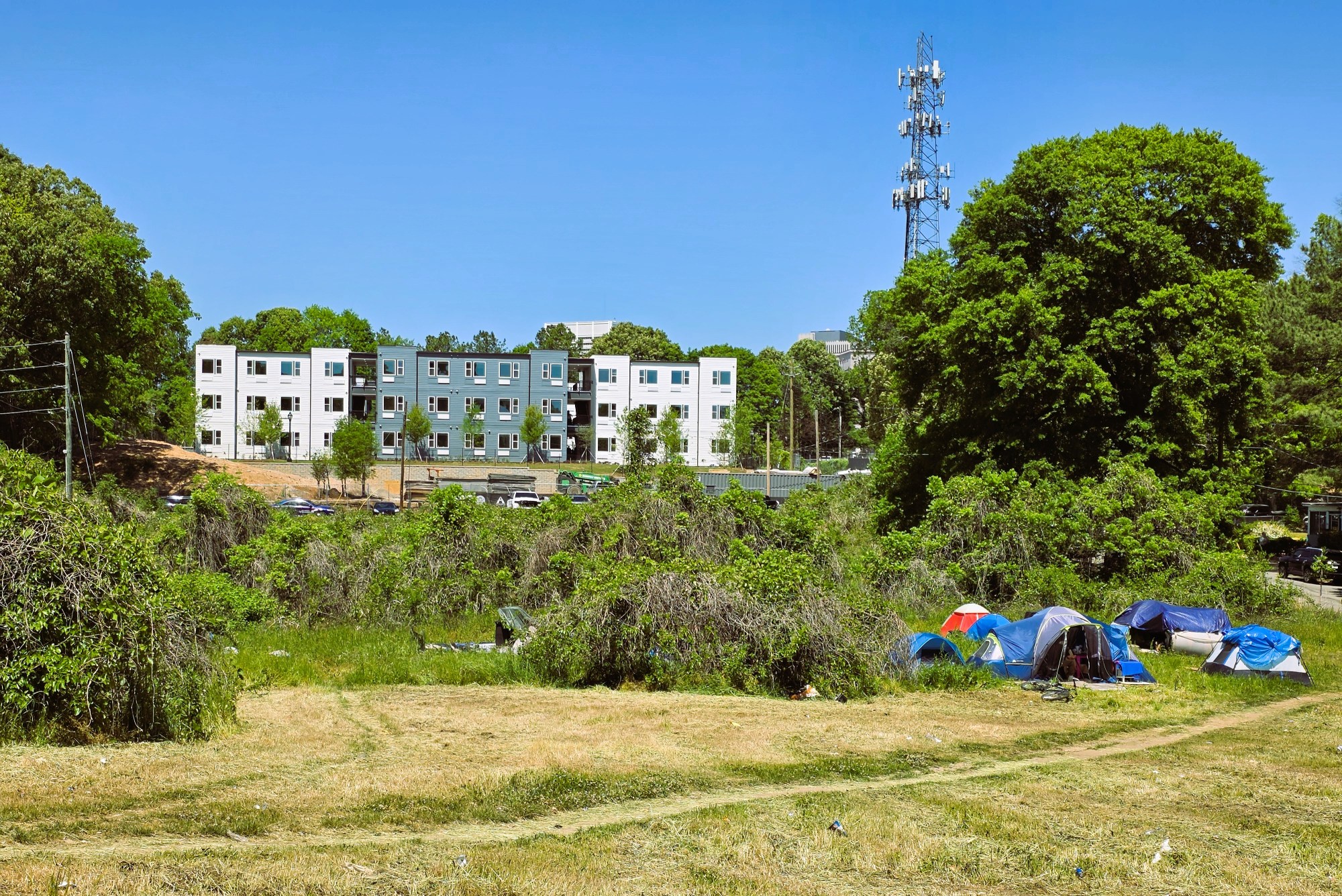 A grey and white apartment complex sits atop a hill. In the foreground, an overgrown lot is dotted with tents, where unhoused people live.