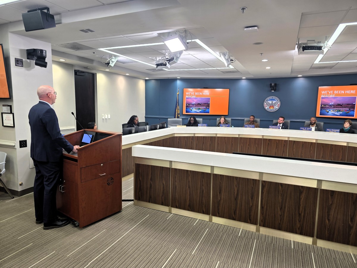 A man in a dark suit stands at a podium, addressing a panel of Atlanta City Council members.